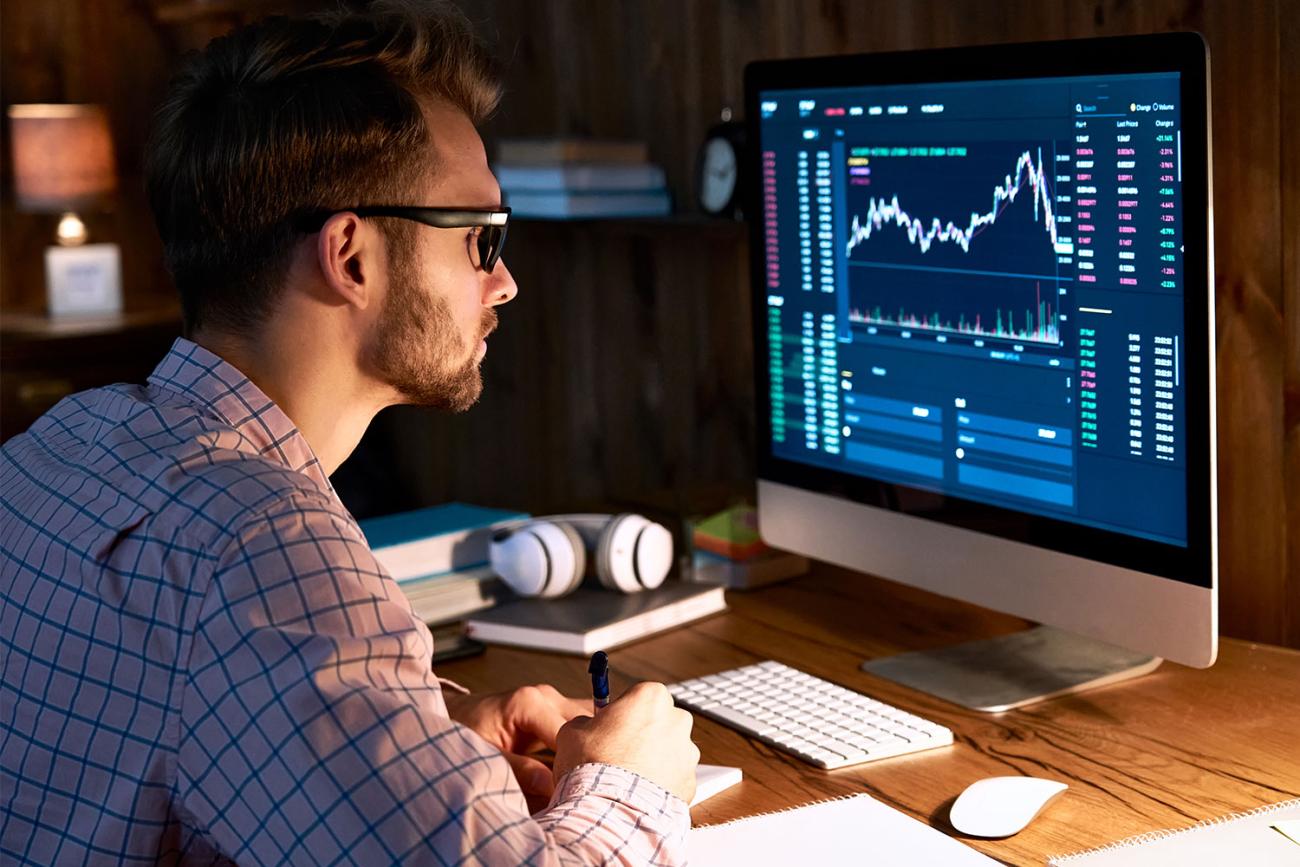 Man with brown hair and black glasses looking at graphs on computer monitor