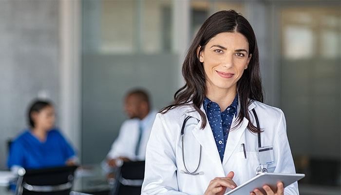 A female nurse practitioner in white coat holding a clipboard in front of a table of colleagues