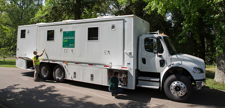 Mobile Civil Infrastructure Laboratory with one person looking in the undercarriage and another, wearing a reflective work vest, inspecting the side wall