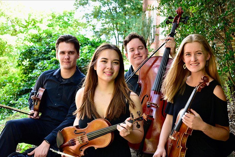 Four people holding various stringed instruments in an outdoor setting