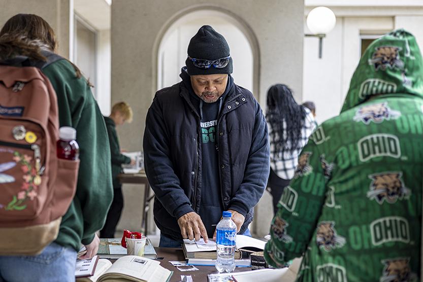 A man in a winter jacket, as well as two people with their backs to the camera, peruse books spread out on a table