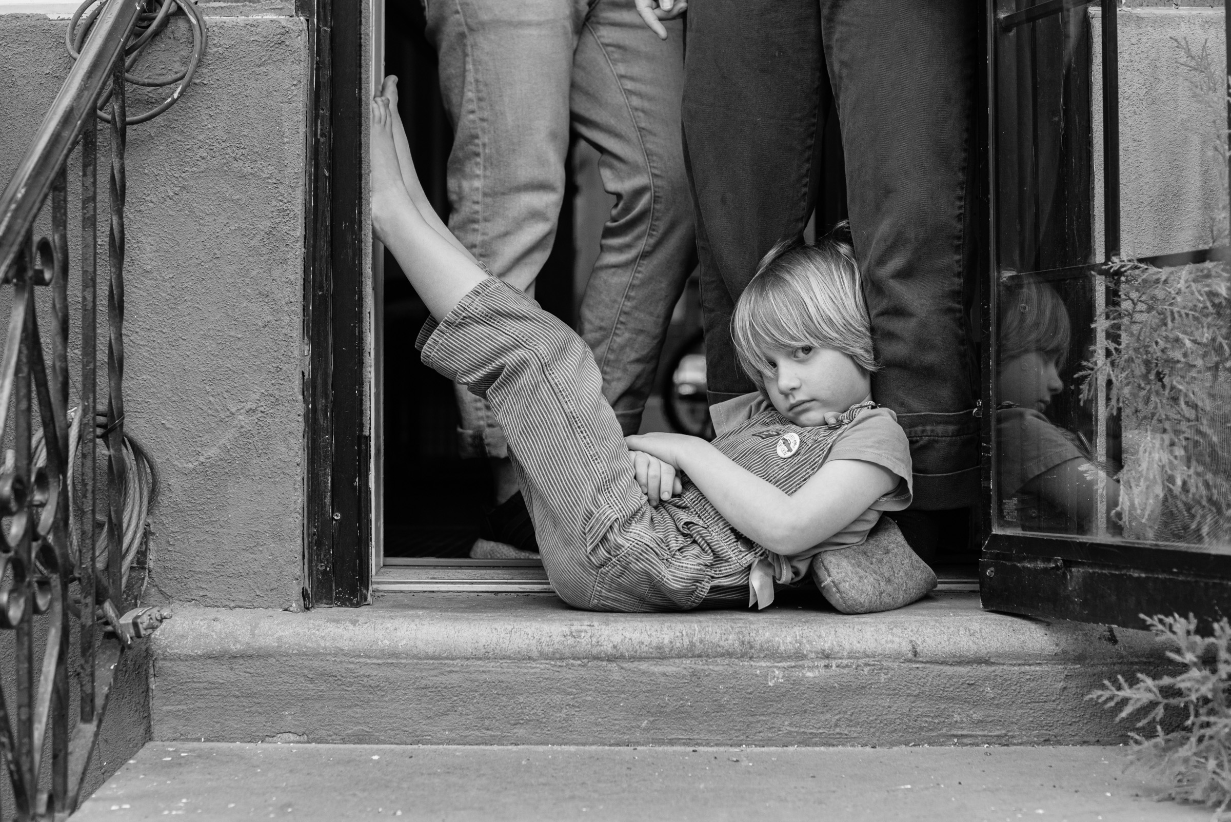 A young child lays in a doorway with his feet up against the doorframe with family members behind him
