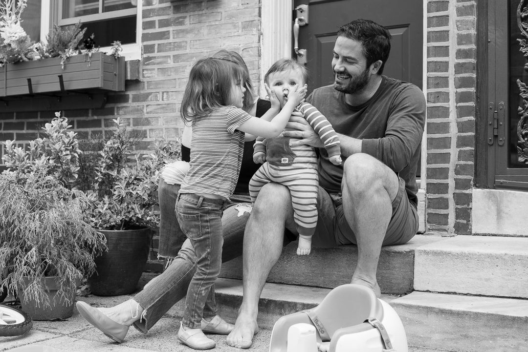Family sitting on their front step laughing at young child playing with toddler