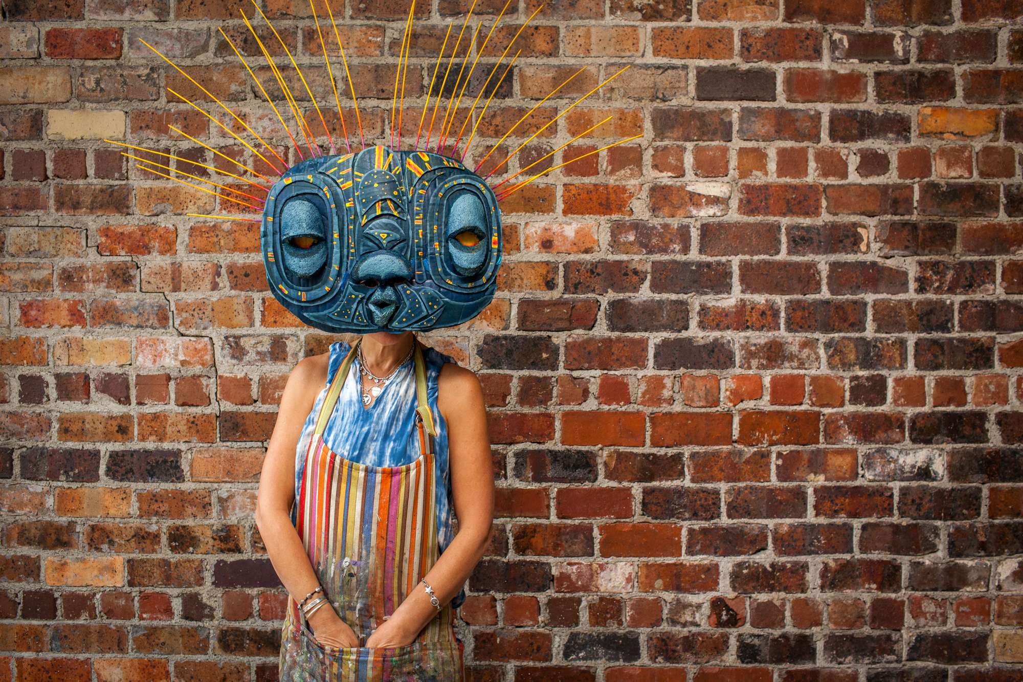 An art student poses against a brick wall wearing an oversize homemade halloween mask that resembles an insect