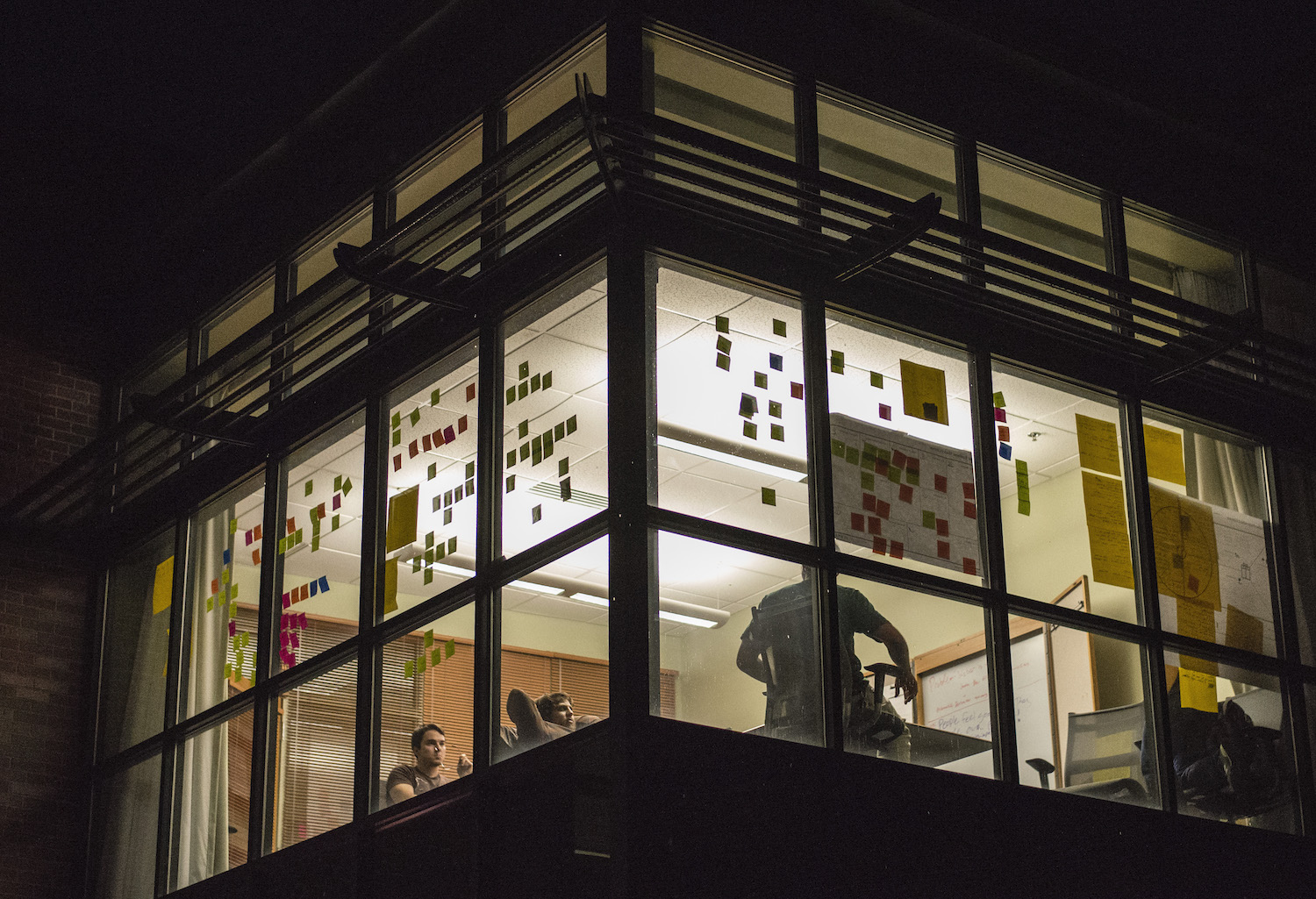 Post-it notes are all around windows of an office as people seem to be working inside, late at night