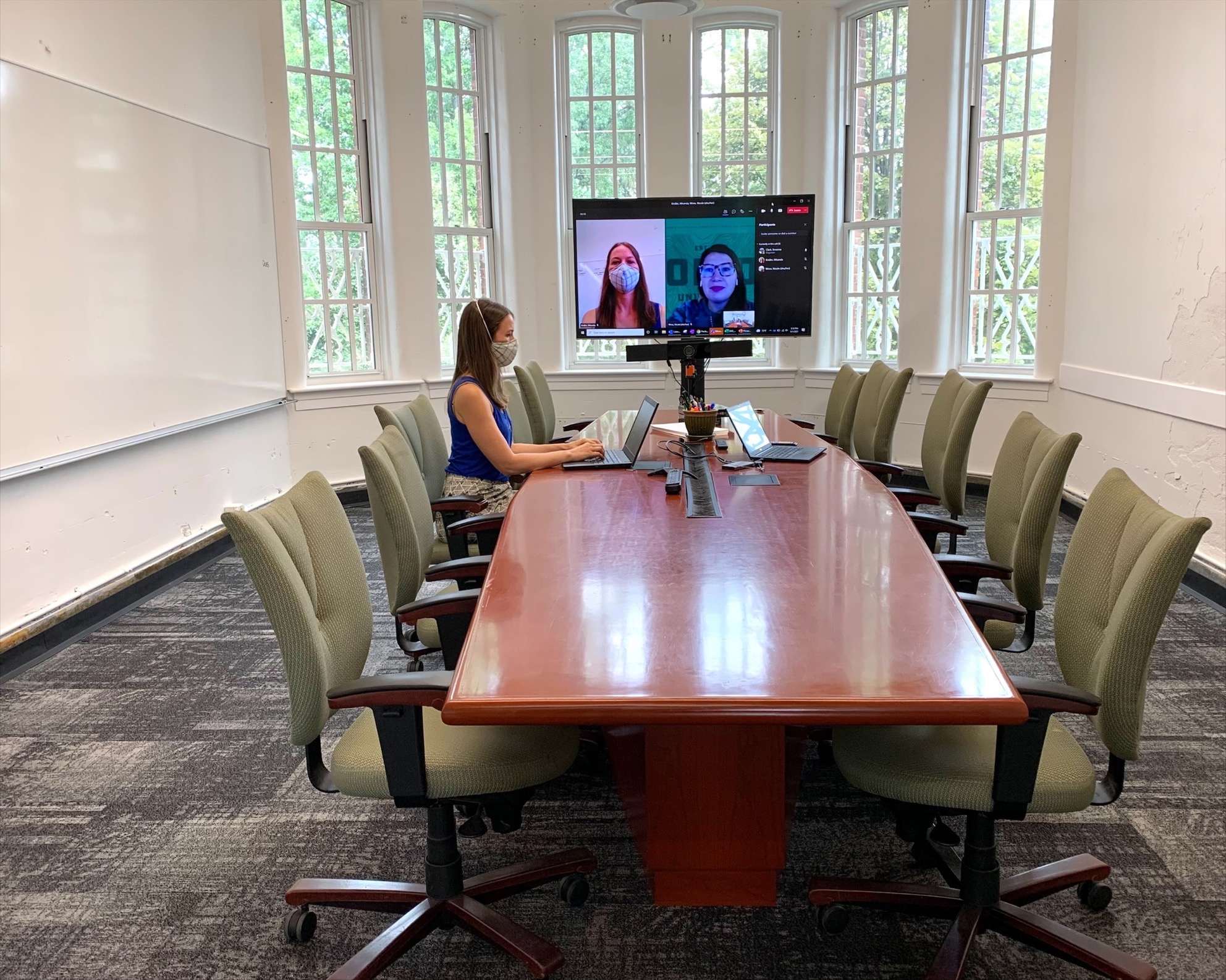 A woman sits at an empty conference table, with two people displayed on the TV using the Teams application