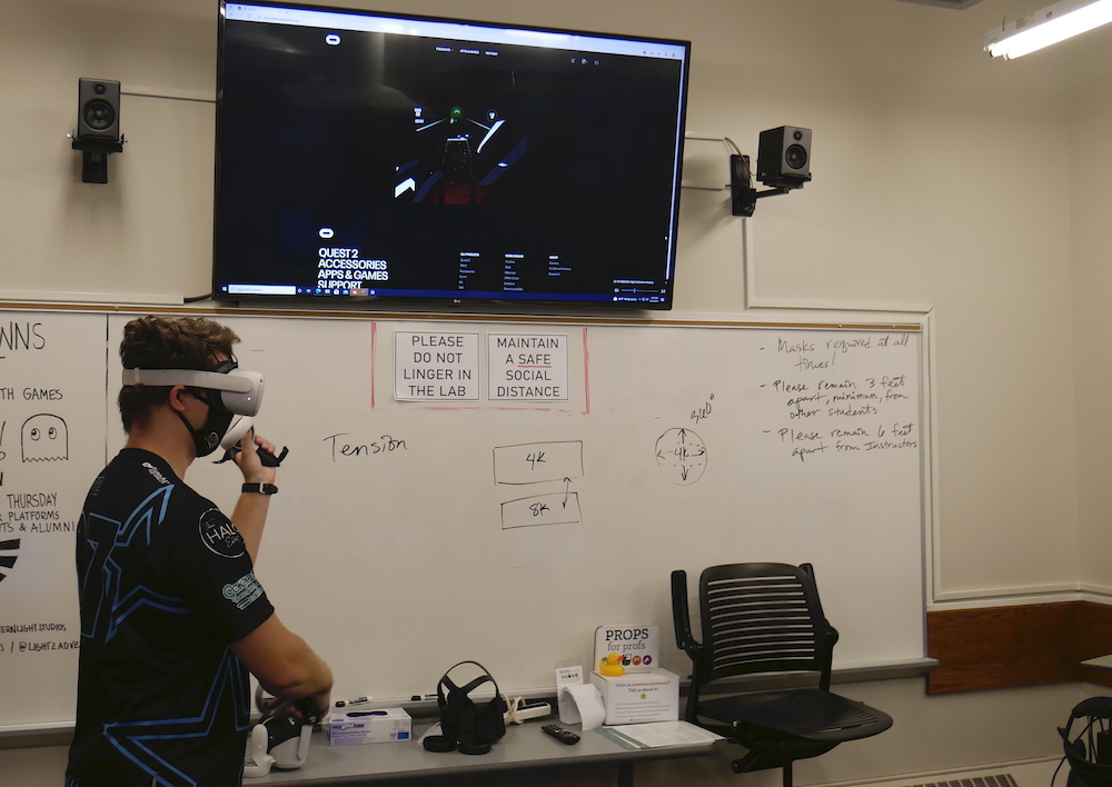 A student stands in a classroom in front of a white board wearing a VR headset. What the student sees in the headset is displayed on a TV that sits above the whiteboard. 