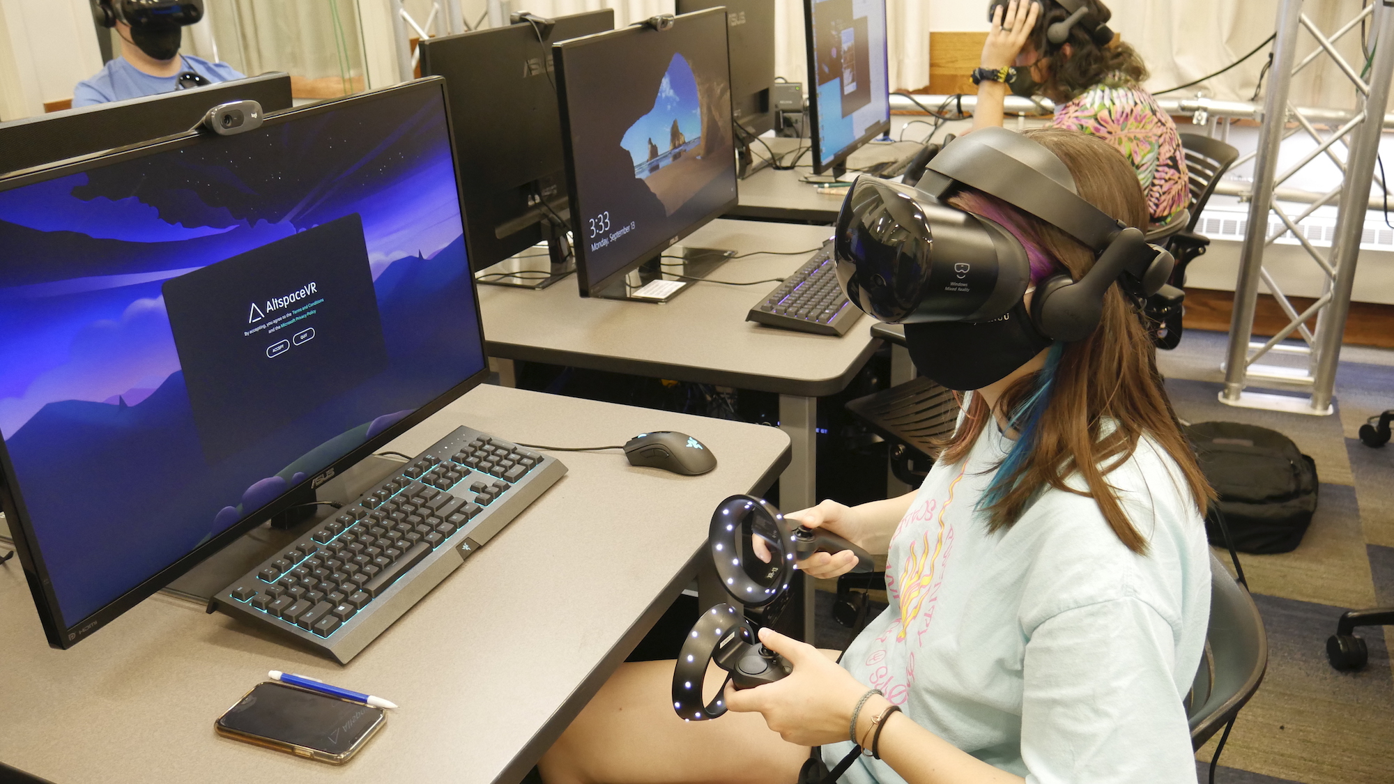 A student with long brown hair sits at a computer desk in a classroom with a virtual reality headset on