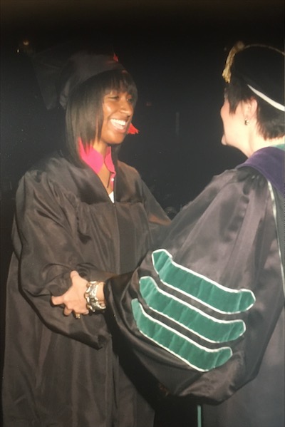 A young woman in a black cap and gown shakes the hand of another person as she is handed her diploma at graduation