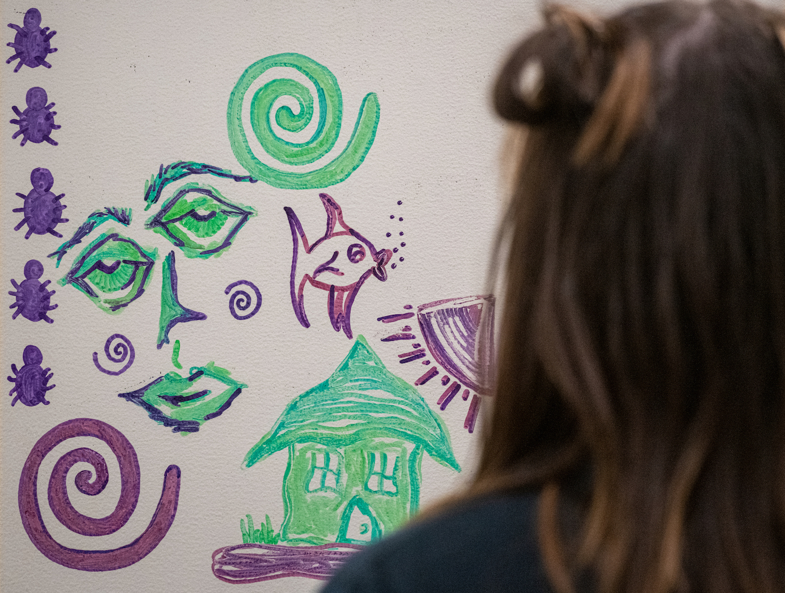 A student has her back to the camera, looking at drawing of purple swirls, spiders, a green face, a green house and more.