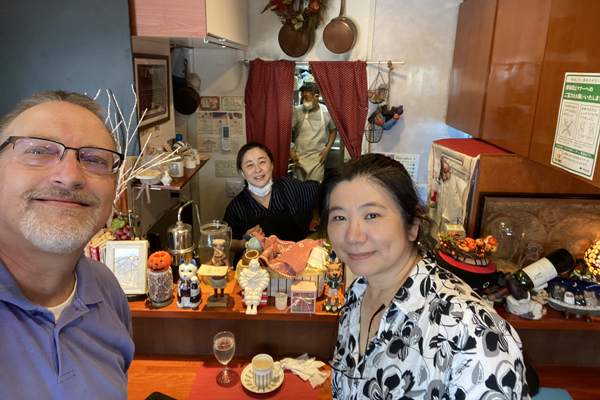 Chris Thompson with Akisa Fukuzawa (Master of Sports Management '85) next to him, Futaba Kaiharazuka (M.A. in Development Studies '00) behind the table, and Nobu Kaiharazuka (B.A of Business Administration '99) with his mask on in the kitchen of Bistro Noubu.