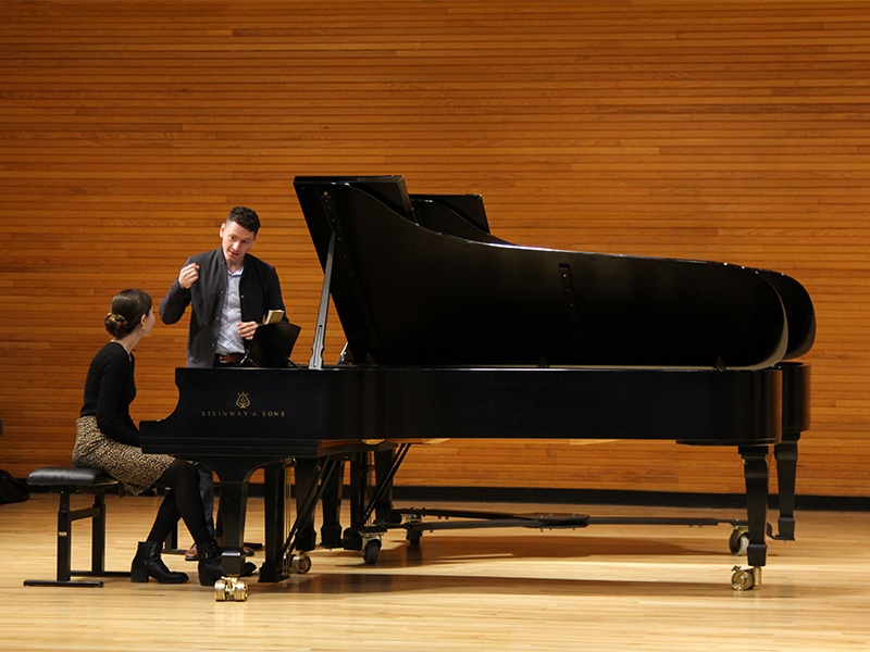 Two people on stage, one sitting at a piano, and the other standing