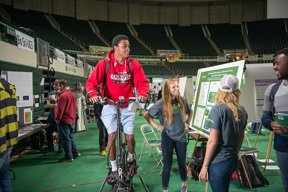 an Ohio University student rides a stationery bike while other students converse around a display at a research expo