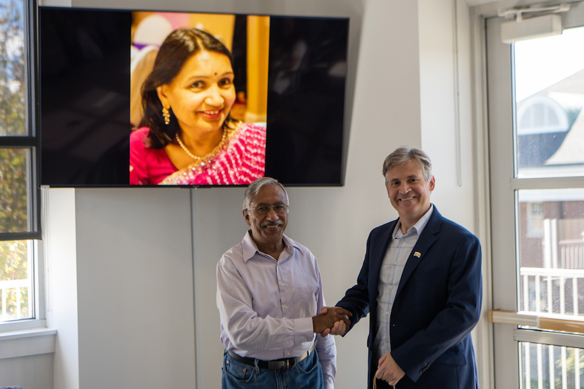 Emeritus professor Ashok Gupta and John McCarthy, Dean of CHSP shaking hands with an image of Sudha Agrawal displayed on TV screen to the left.