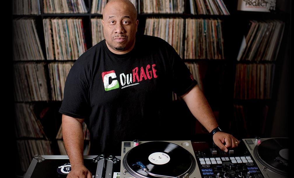 Jason Rawls in front of a turn table, looking up at the camera, wearing a shirt that says 'Courage' in white, green and red.