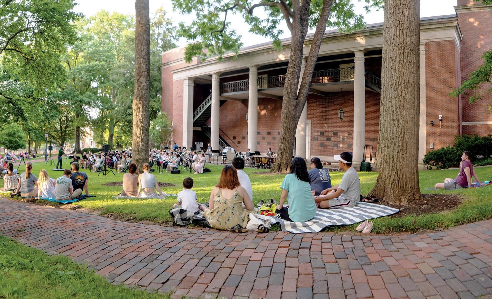 People sit on the grass near Memorial Auditorium, listening to an orchestra playing on College Green.
