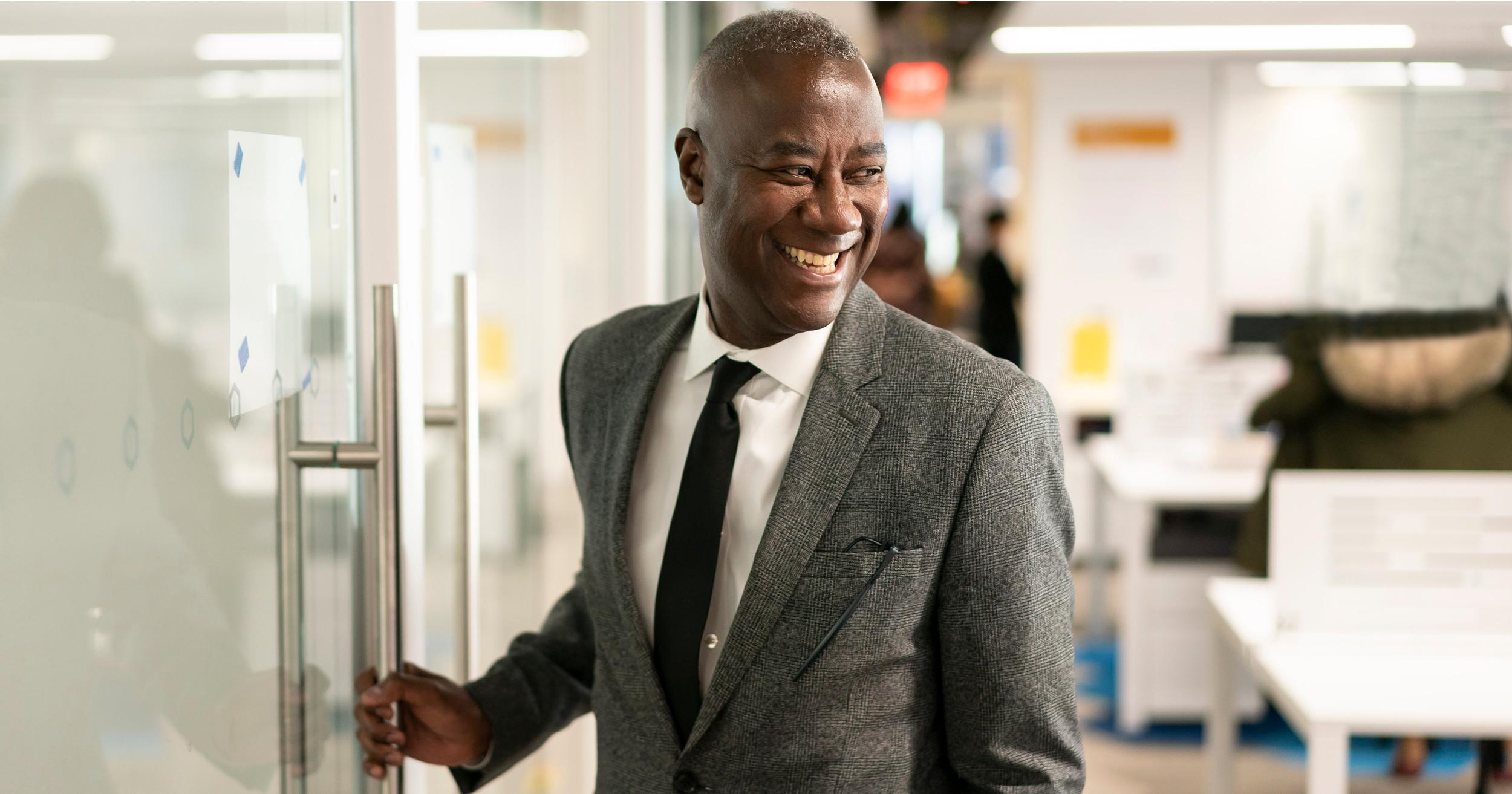 Byron Ward stands in a grey suit, smiling and looking back as he opens a door.