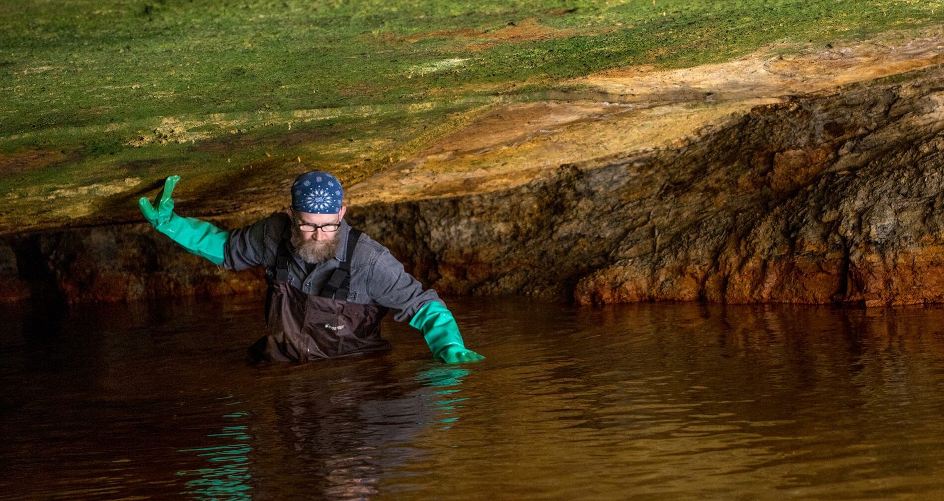 A man stands in a cave, waist-high in water, with a hand reaching up and touching the low cave ceiling near his head.