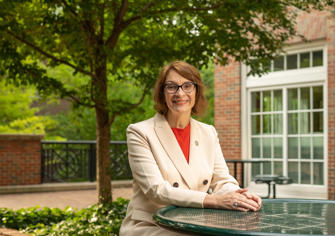 Professor Kerri Shaw poses at an outdoor table wearing an blazer and eyeglasses