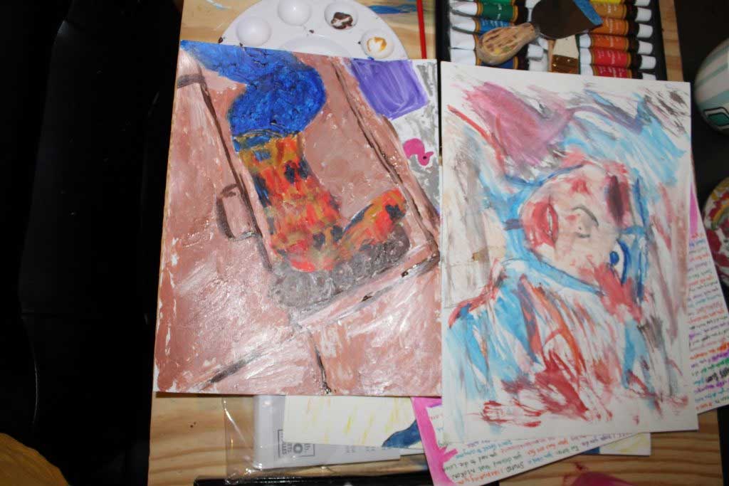 Two paintings lay upon a paint palette, a set of acrylic paints, artist tools, and other barely seen paintings. The paintings are horizontal and are placed one above the other. The painting near the top of the photograph is made with paint of varying blues and reds, merging to brown in some areas. The background is made in streaks of blue and red. In the middle is a person’s face, neck, and shoulders, painted in the same streaky style as the background. One eye is open, looking down, with bruising to the le