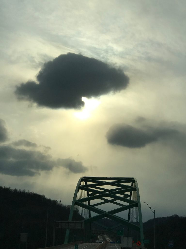 The sun peaks through a cloud in a gray sky, with the top of a bridge stretched down below it. There are cars moving on the bridge.