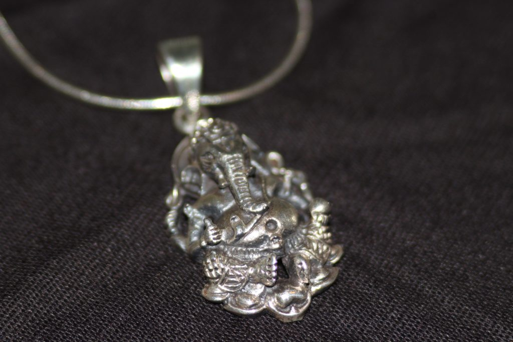 A silver necklace of Ganesha, God of new beginnings, success and wisdom, and Remover of Obstacles, lays on top of a dark knit background. The bottom of the Ganesha pendant is in focus, and the image blurs as it moves up Ganehsa and to the necklace.