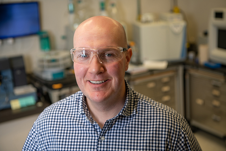 Jason Trembly smiles at the camera, he is standing in a lab with safety goggles and wearing a blue plaid shirt.