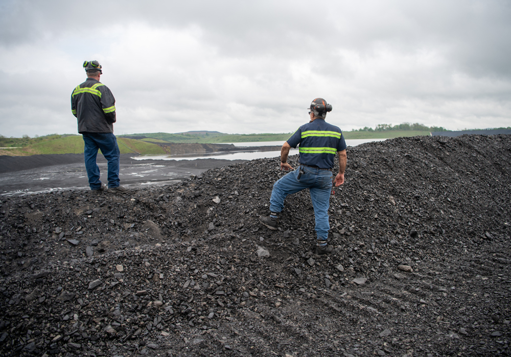 two men in high-visibility shirts stand on a mound of coal