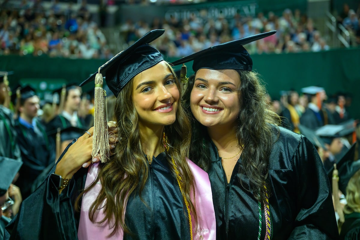 Two female graduating students sit side-by-side and take a selfie using a phone