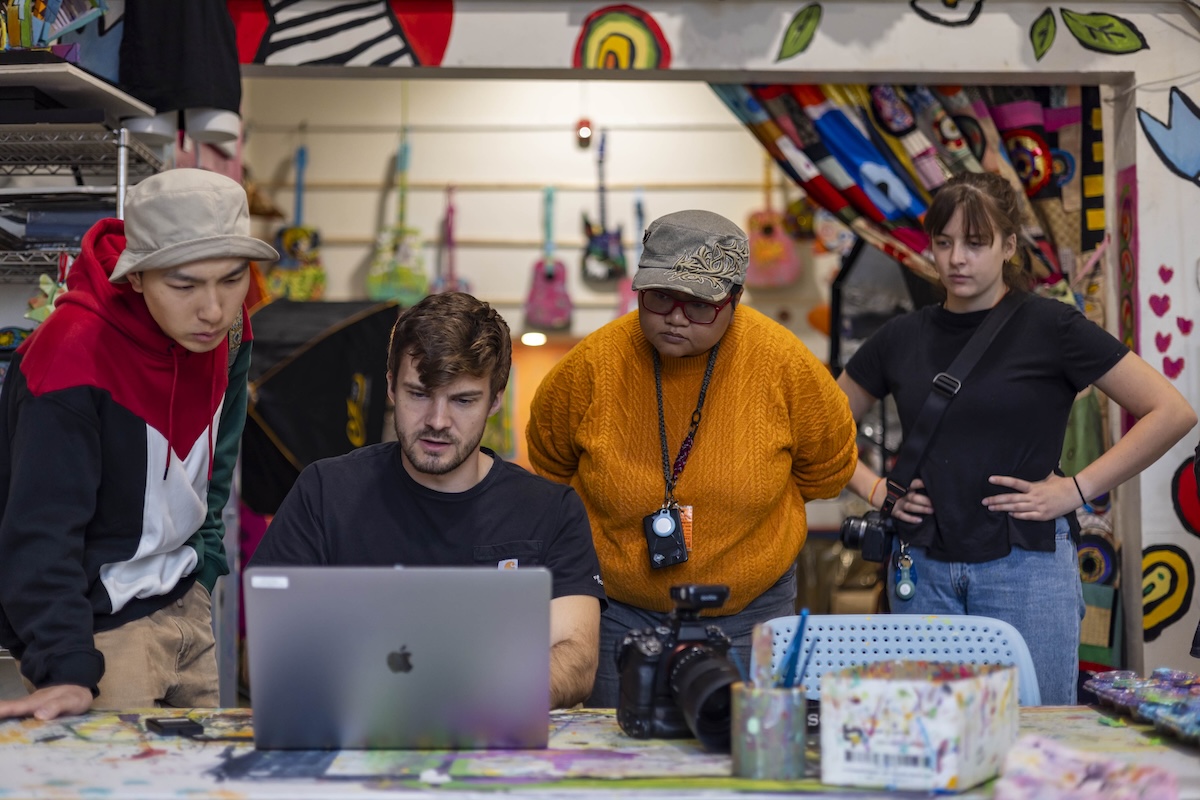 Three people look over the shoulder of a seated man who is typing on a laptop.They are in an art studio.