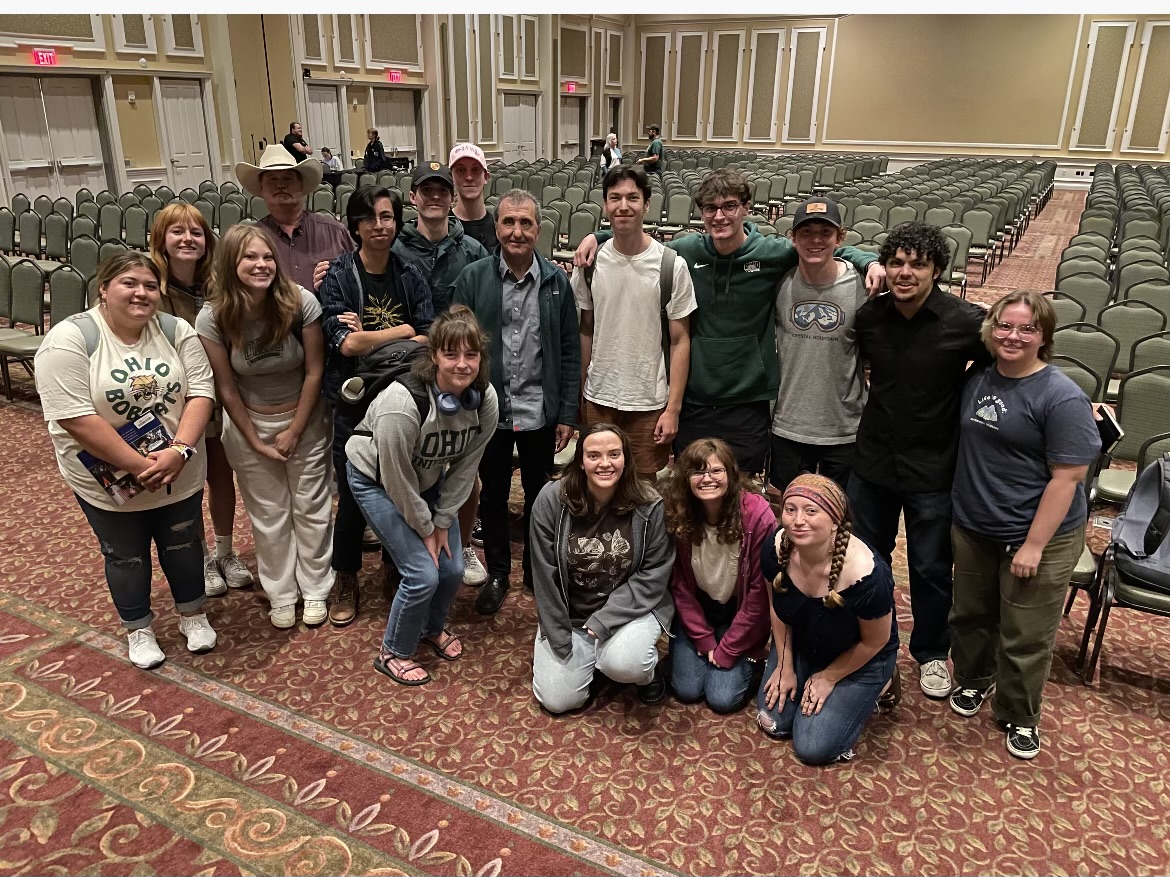 A large group of students and professors pose together in a large theater setting, smiling at the camera