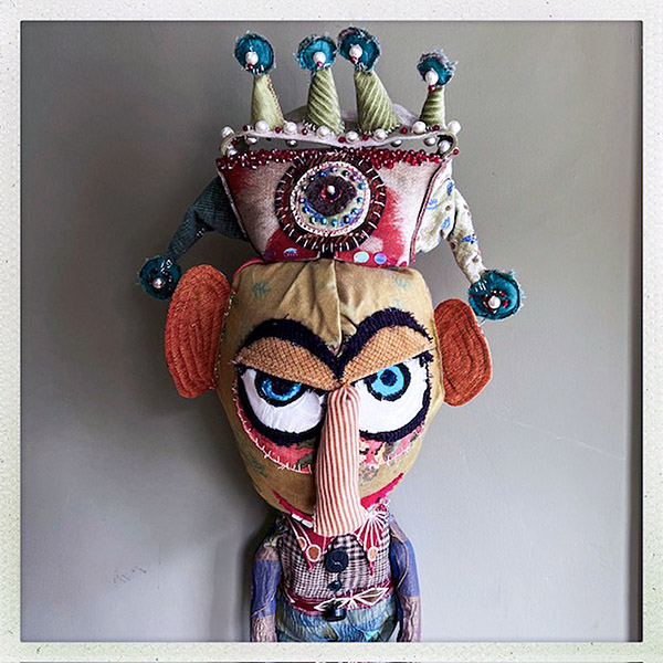 a fabric puppet wearing a multicolored crown and clothing with blue eyes, large red ears and a long red and white striped nose