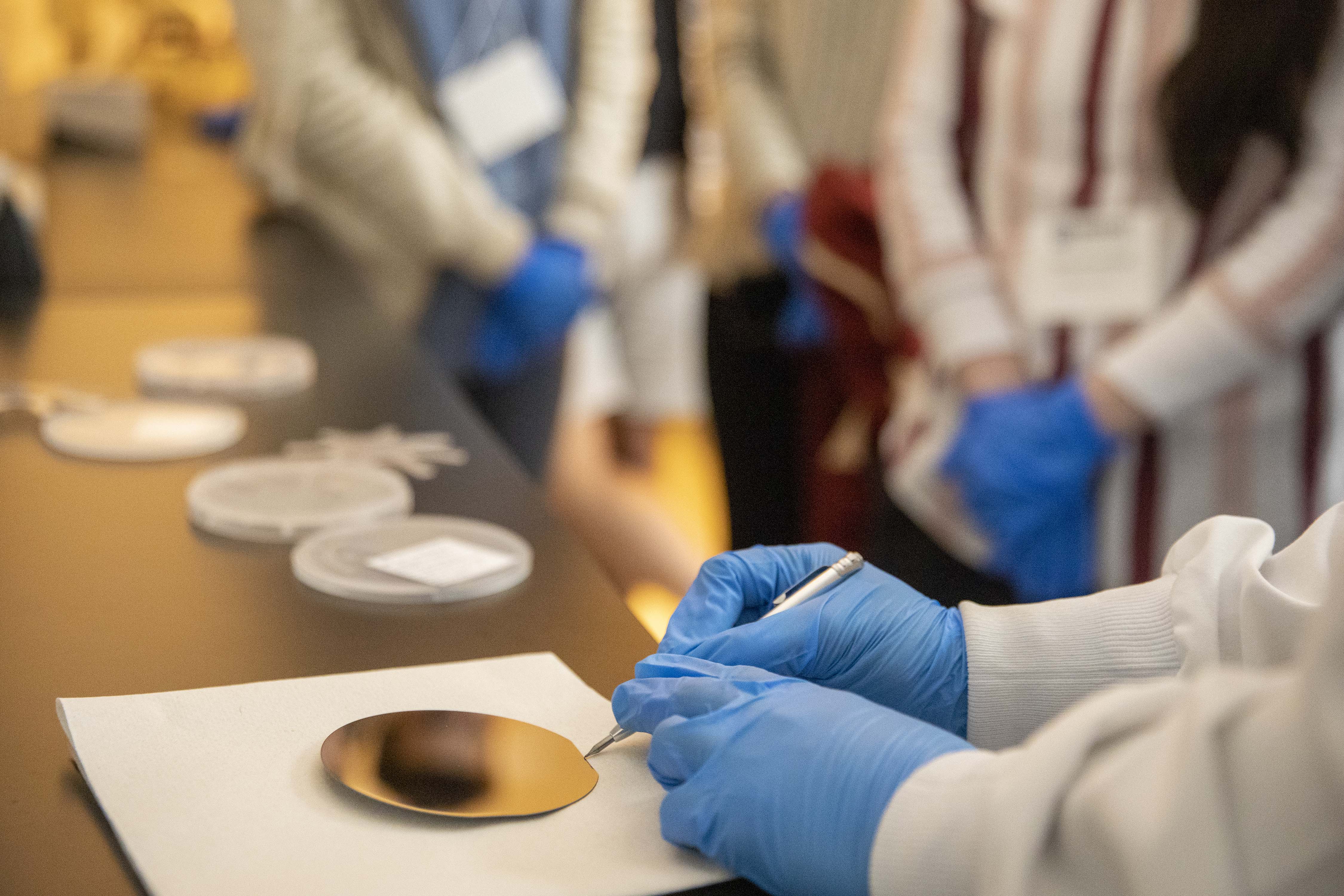 a pair of hands wearing nitrile gloves and a lab coat works uses a metal tool to point to a circular metal semiconductor piece