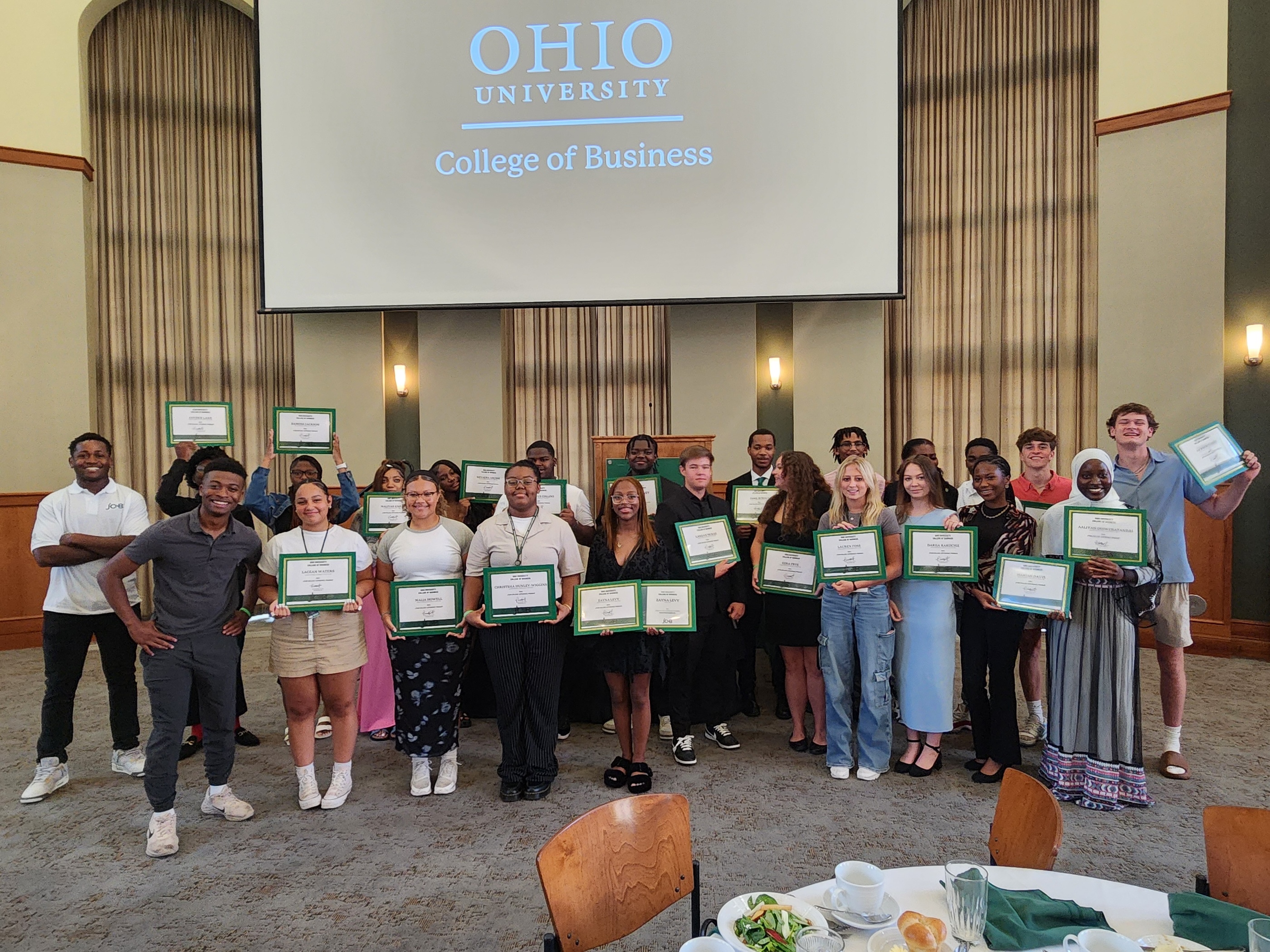 A group of students stand in a large reception room posing for a picture, holding certificates