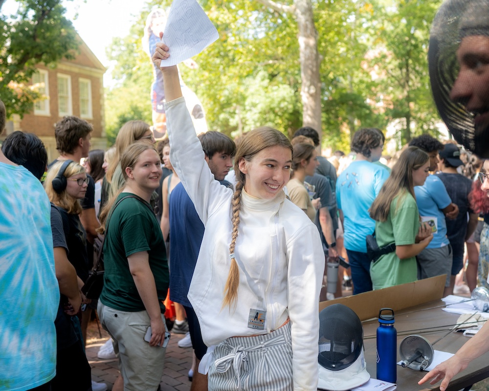 A student wearing a fencing uniform holds up a stack of fliers