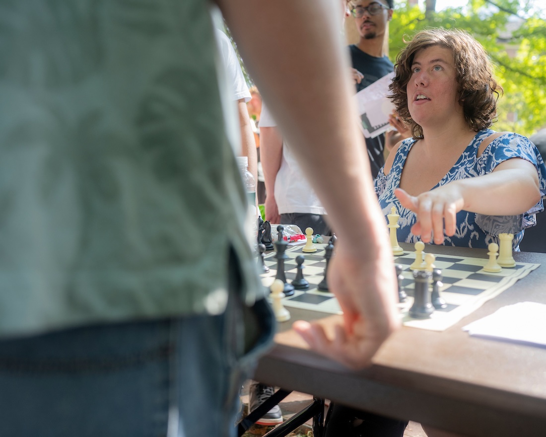 A woman sits in front of a chess board, talking to someone standing in front of her whose back is to the camera