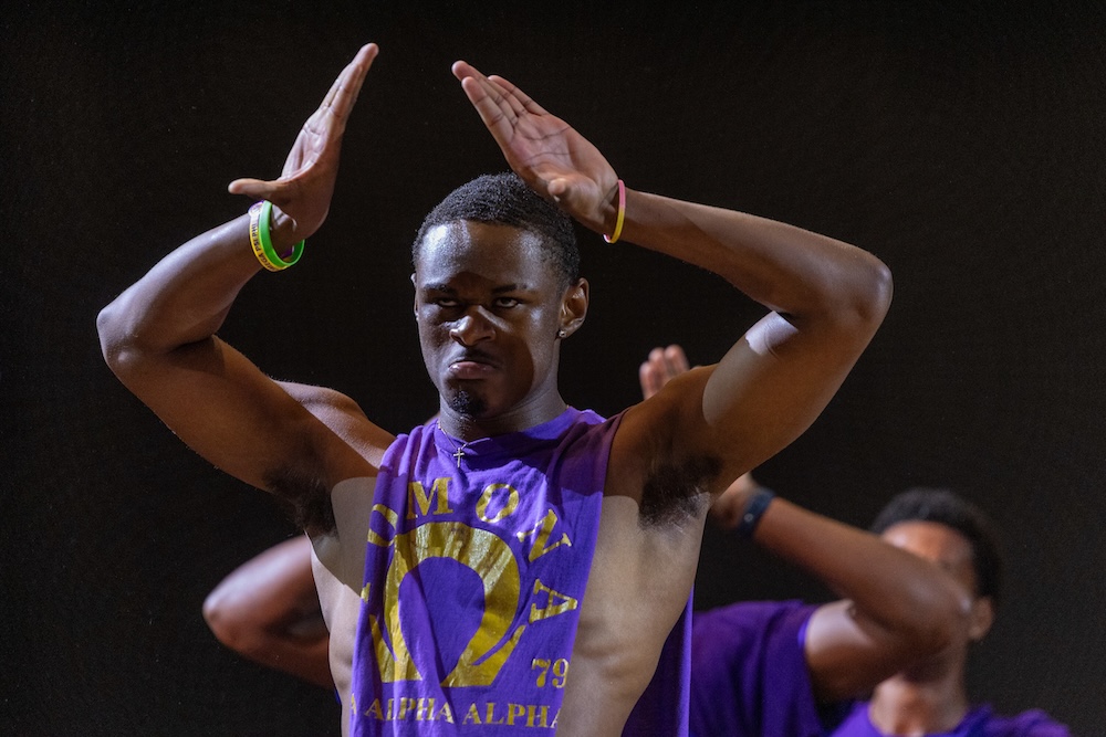 A student wearing a purple fraternity shirt performs, his hands raised over his head