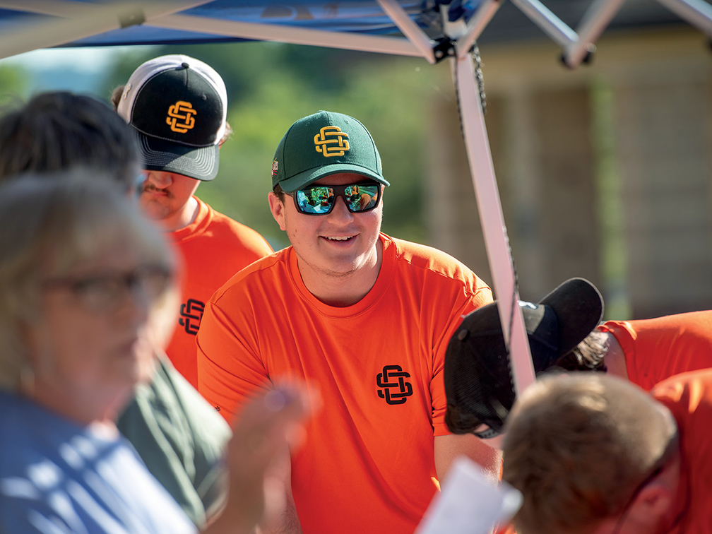 a student in an orange shirt, green hat and sunglasses stands under a sun umbrella