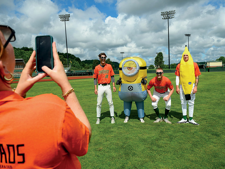 a woman in an orange shirt uses her cell phone to take a photo of two Copperheads players, a person in a banana costume and a person in a Minions costume on the outfield at Bob Wren Stadium