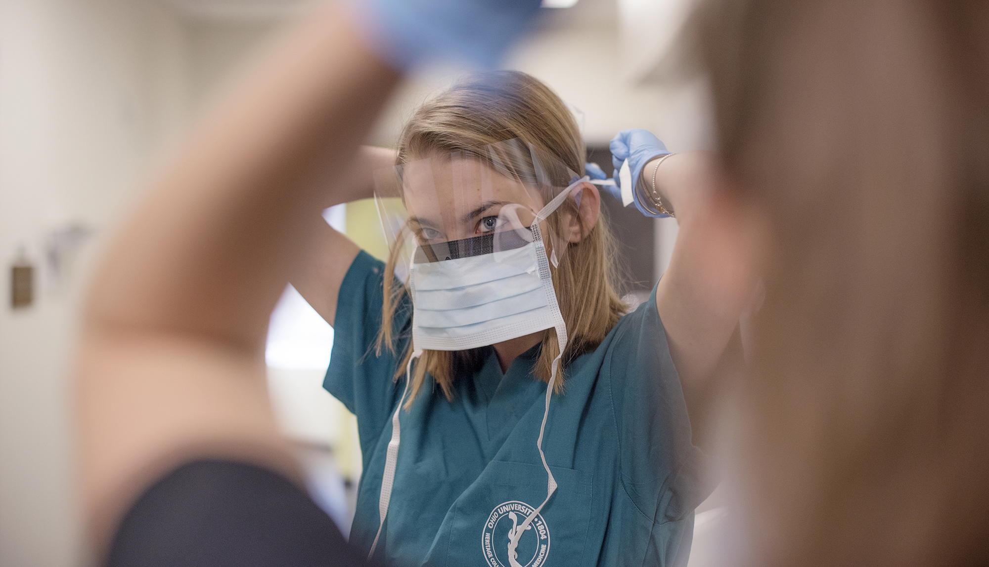 a female student wearing scrubs puts on a disposable face mask and plastic eye shield