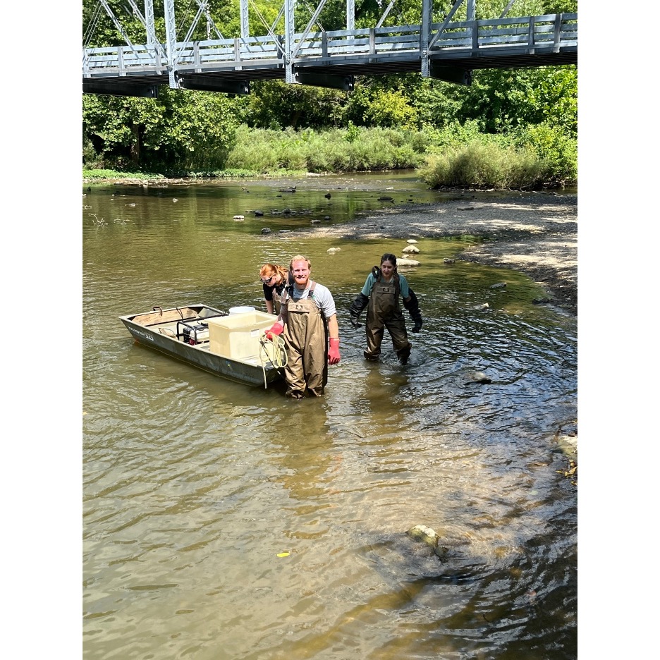 People in fishing gear stand knee-deep in a stream by a small boat