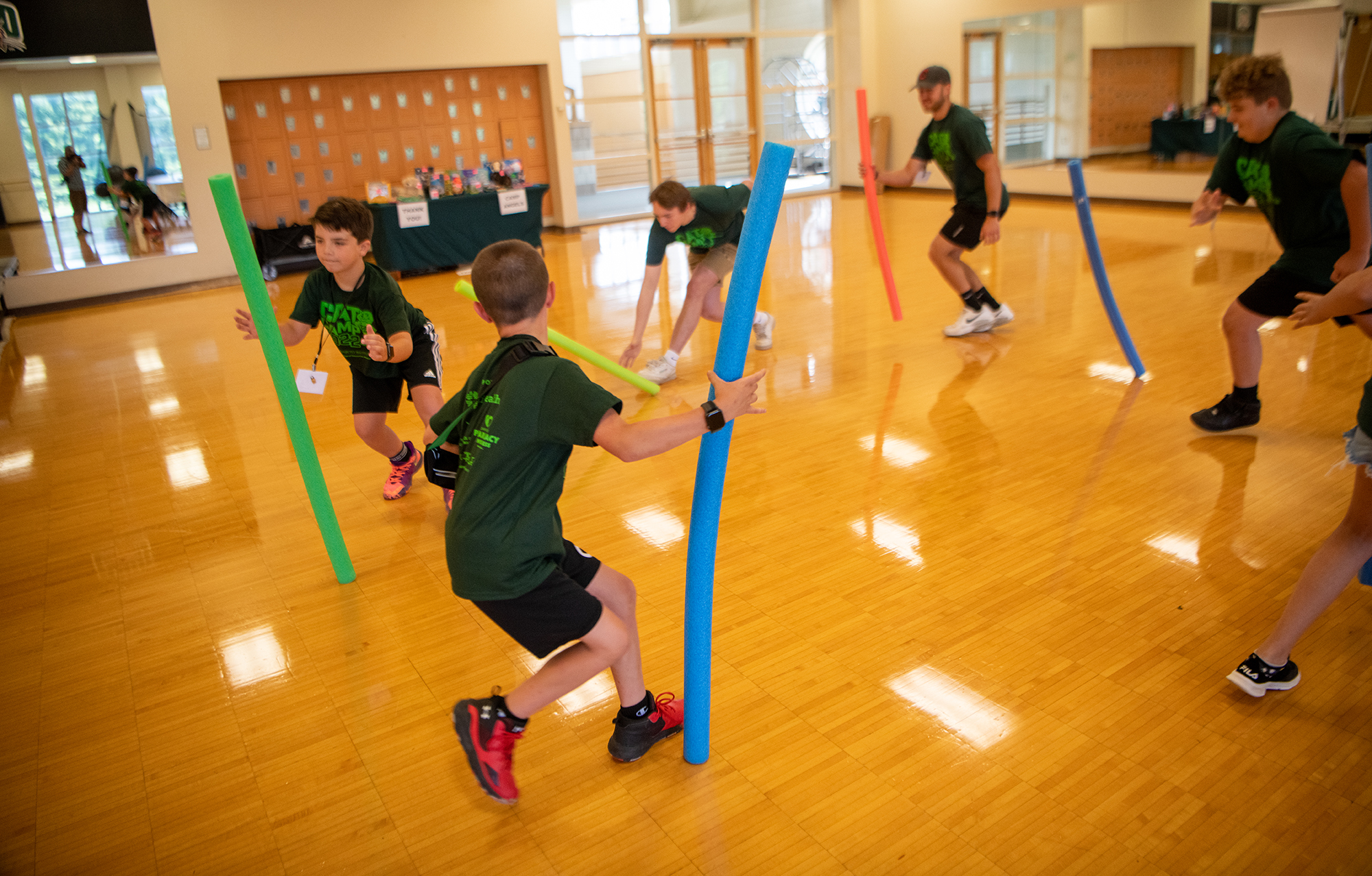 : two young boys and two older camp counselors play with pool noodles in a gymnasium-style room