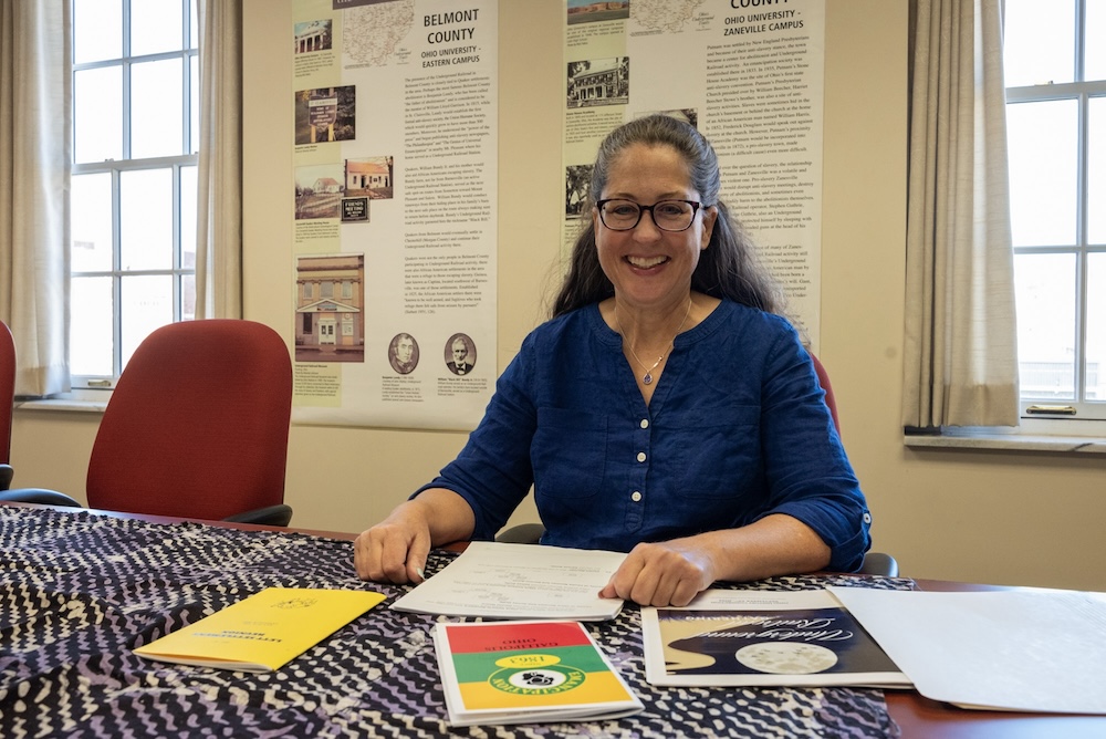 A woman sitting at a large table with documents in front of her smiles at the camera