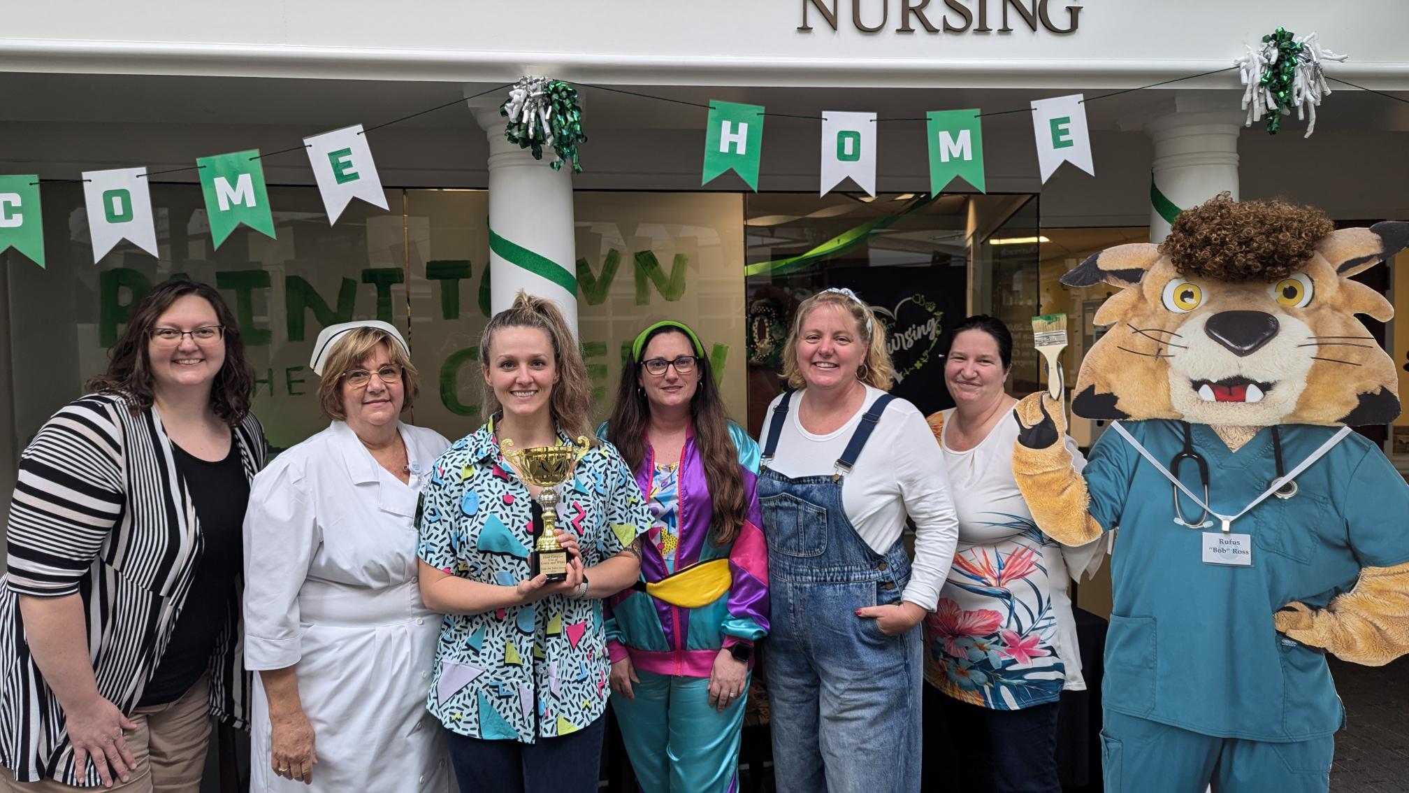 Faculty and staff from the School of Nursing pose with an image of Rufus dressed as a nurse, in an office decorated for Homecoming