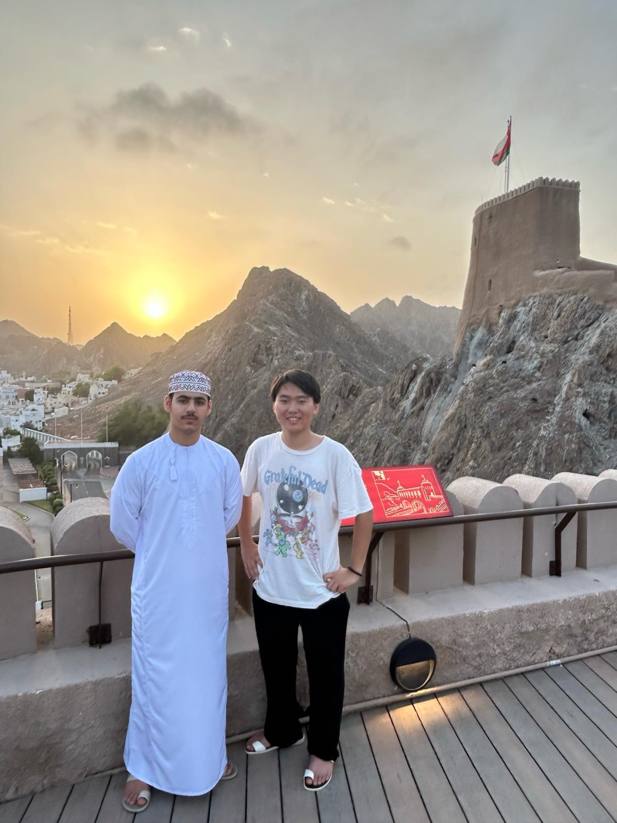 Two men stand side-by-side in front of a railing, the sun setting behind them