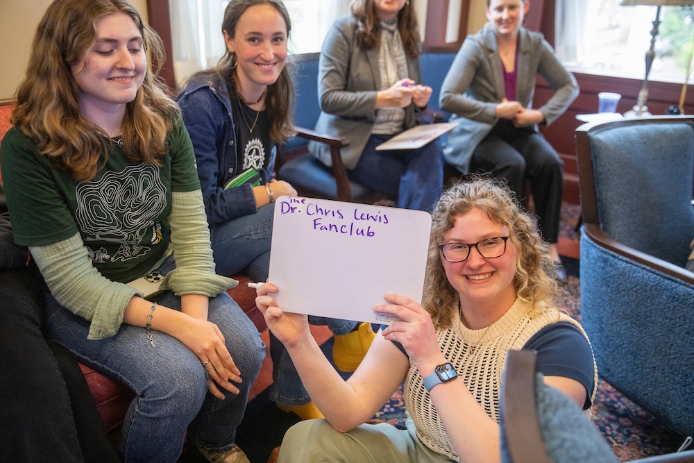 A group of college students poses, smiling. One is holding a board reading 