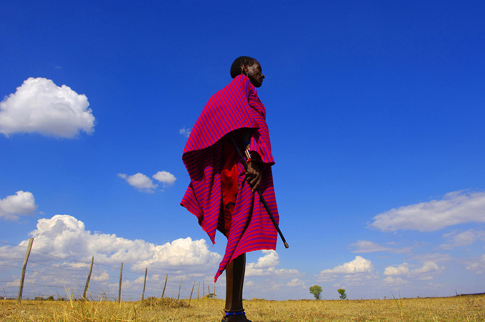 a black man in a red and blue shawl stands in an open field under a blue sky