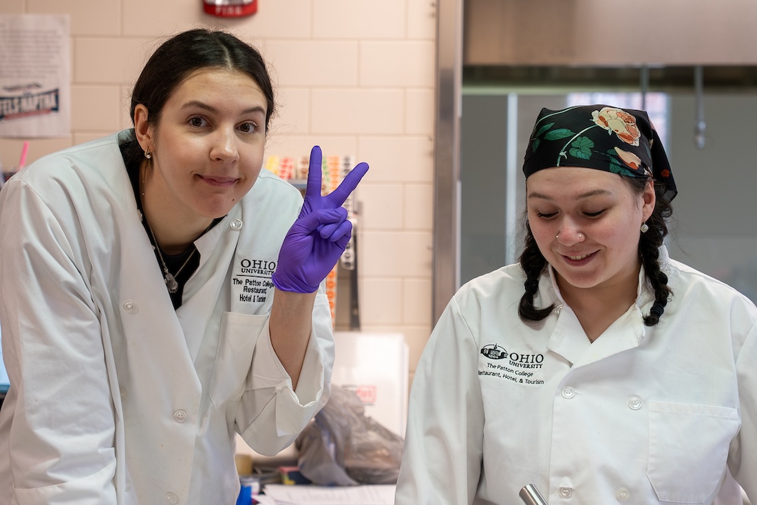 Two students wearing white chef's jackets stand in a kitchen. The one of the left is wearing purple gloves and flashing a peace sign.