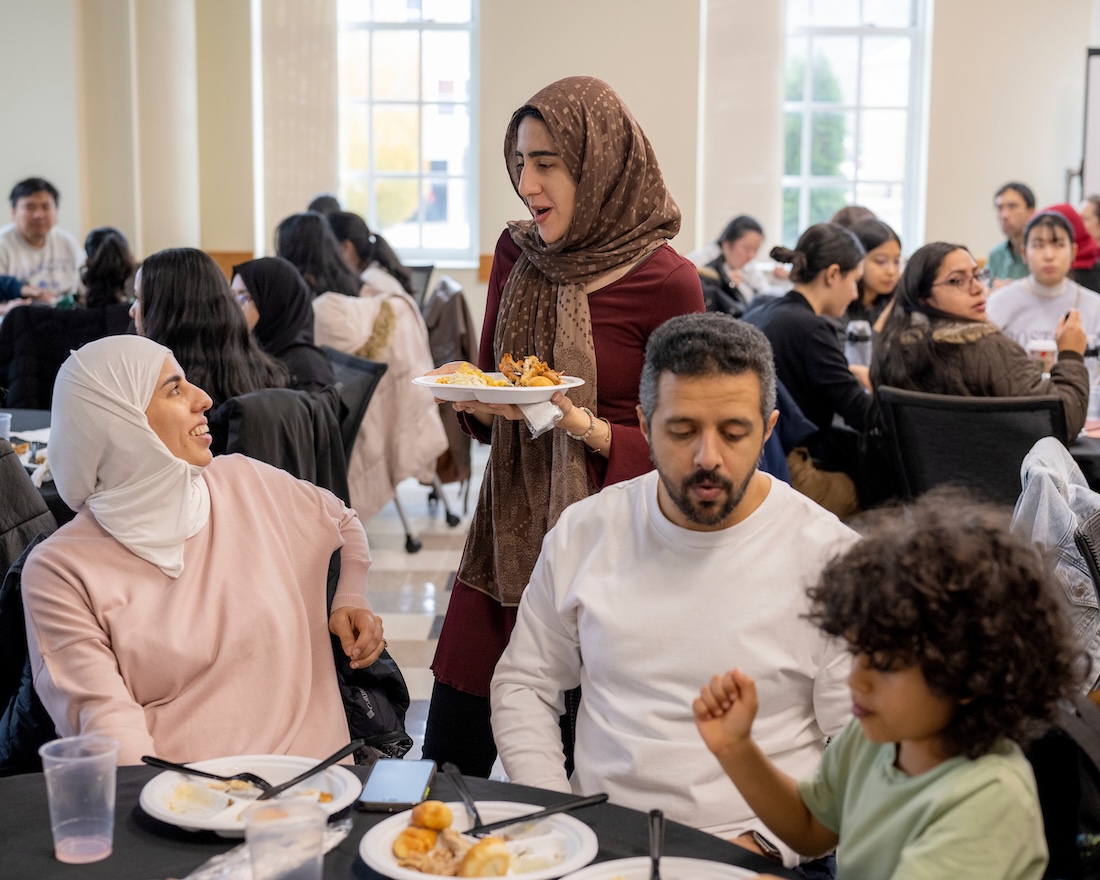 In a crowded room filled with tables, two women (one seated, one standing) talk to each other