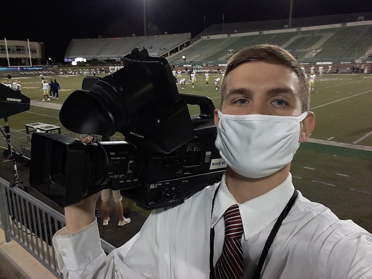 Bryan Kurp in a selfie wearing a face mask and holding a large video camera on his shoulder, with an Ohio University football game in the background. The stands are empty.